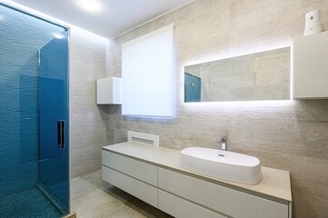 Modern bathroom with blue glass shower, sleek vanity, and illuminated mirror. Neutral tones create a clean, minimalist aesthetic
