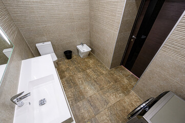 Modern bathroom. Neutral tiles cover walls and floor. Features a white sink, toilet, and bidet. Dark wooden door is seen