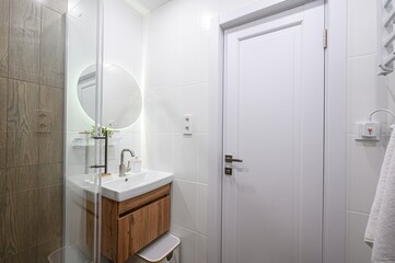 modern bathroom with a wooden vanity, sink, round mirror, and white door. The walls are tiled, creating a clean, minimalist aesthetic