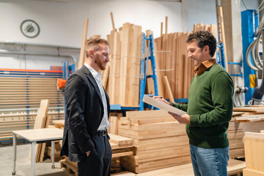 Two carpenters talking in front of stacked wood planks in production hall