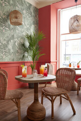 A vibrant tropical-themed restaurant interior featuring colorful drinks on a round table, surrounded by wicker chairs and lush green wallpaper
