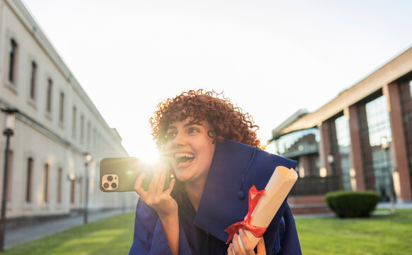 Young woman celebrating graduation on university campus holding diploma and phone