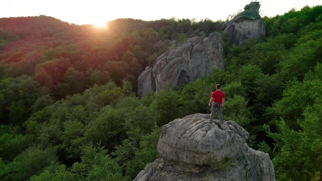 Aerial view of climber standing atop rock formation at Dovbush Rocks in Carpathian mountains, Ukraine. Sun sets, casting warm glow over lush, green forest, distant hills, creating breathtaking view.