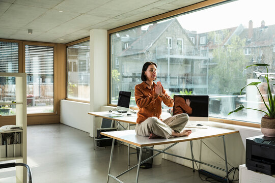 Mature businesswoman in lotus position meditating with prayer pose on desk at office