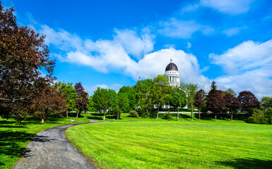 In Augusta, United States, the Maine State House dome peeks above leafy Capitol Park, where a curved gravel path winds across bright spring lawns under a vivid blue sky.