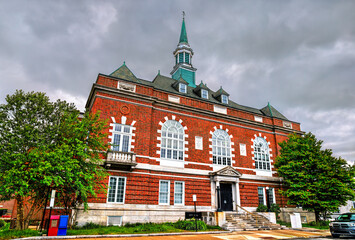 Concord City Hall in New Hampshire, United States, shows its red brick facade, tall arched windows and copper spire beneath dramatic gray clouds and lush spring trees.