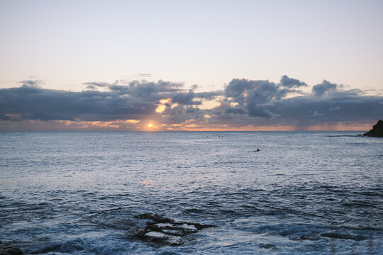 Sun rising over ocean with cloudy sky and surfers waiting for wave