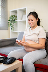 Obraz premium Pregnant asian woman with bare belly drawing her blood for a glucose testing kit as sitting on sofa.