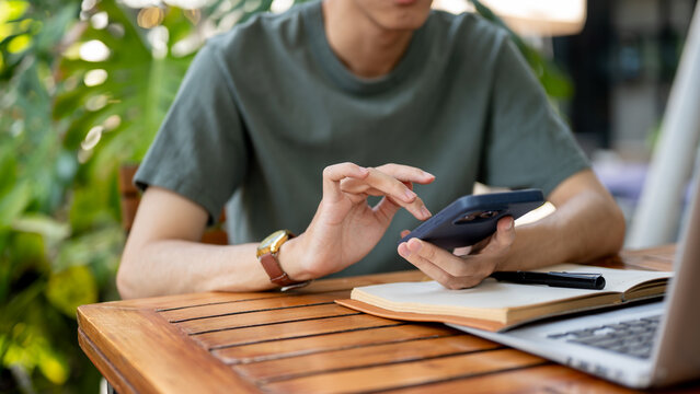 Close up of a man typing on smartphone holding in a hand over a notebook and sitting at wooden table