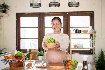 Pregnant asian woman smiling as she showing a fresh vegetable as food ingredients in the kitchen.