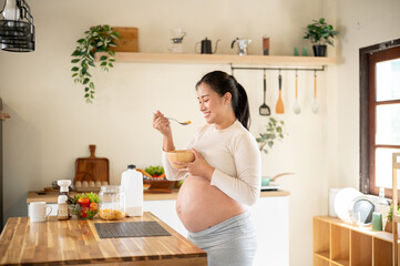 Pregnant asian woman holding a wooden bowl enjoy eating a cereal while standing at kitchen counter.