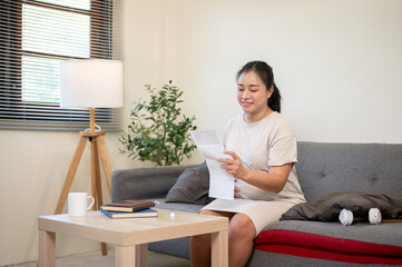 Pregnant asian woman is looking happily at fetal ultrasound pictures while sitting on sofa in house.