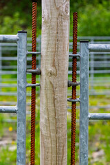 Close-up of rusty gate hinges connecting to a wooden post in a green rural setting, showcasing weathered materials and nature.
