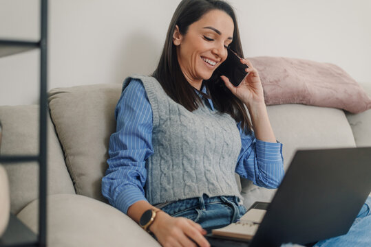 Smiling freelancer working from home on sofa with laptop and phone - Powered by Adobe