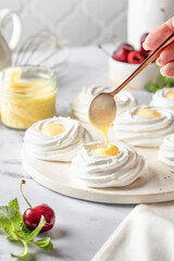 Meringues mini pavlova cakes on a tray on white marble background, process of filling with lemon cream. Light summer dessert