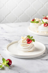 Meringues mini pavlova cake with whipped cream, fresh cherry and mint leaves on a plate, white marble background. Light summer dessert, copy space