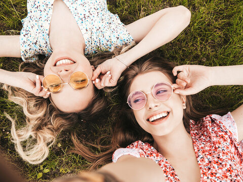 Two young beautiful smiling hipster female in trendy summer sundress.Sexy carefree women lying on the green grass in sunglasses.Positive models having fun.Top view.Taking selfie photos on smartphone