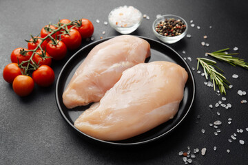 Raw chicken breasts on black plate with tomatoes and spices on dark background