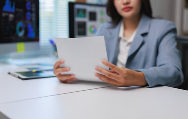 Businesswoman reading documents at office desk with charts on computer screen