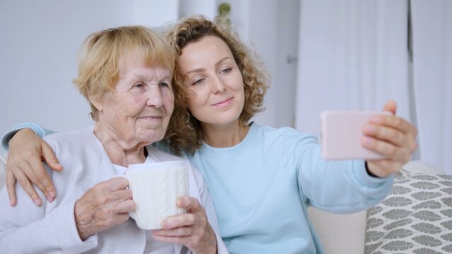 Young nurse embracing elderly woman while enjoying a warm beverage from a mug, capturing a joyful selfie with a smartphone in a cozy nursing home setting
