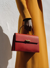 Close up of a fashion model holding an elegant red leather handbag while wearing a stylish mustard yellow coat, showcasing a contemporary and sophisticated look