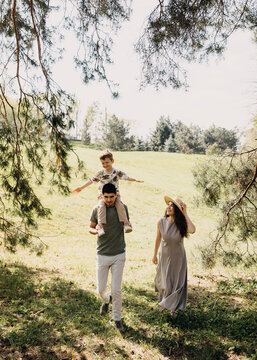 Young family walking through a sunny meadow, father carrying son on shoulders while mother walks beside them smiling.