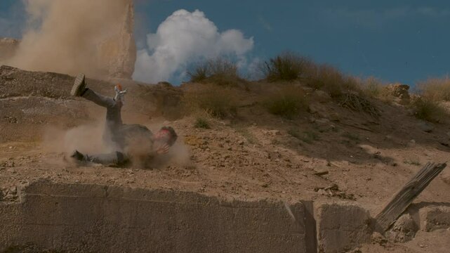 Man with toy gun stumbling  down earth mound as explosion goes off in super slow motion