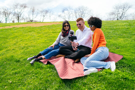 Three content creators sitting on blanket in park, filming video podcast using smartphone with microphone