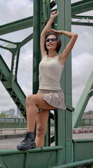 Obraz premium Stylish young Asian woman poses on a green metal bridge in an urban setting. She wears a beige sleeveless tank top, a short pleated khaki skirt and black combat boots, exuding a trendy and modern vibe