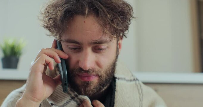 Sick young man talking on the phone with doctor while sitting on the sofa, sneezing and caughing as having a cold. Concept of healthcare and illness.