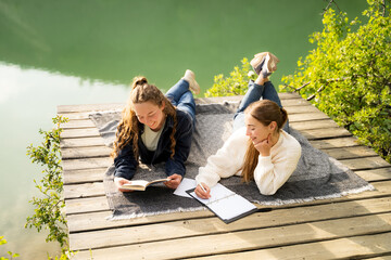 Two young women studying and reading on wooden deck by mountain lake wear casual lying on a picnic blanket, outdoor education and friendship, spring lifestyle, countryside leisure activity in nature
