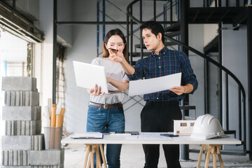 Asian couple reviews architectural plans together at renovation site. They collaborate on ideas to...