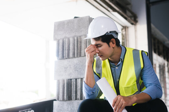 Asian male engineer wearing a hard hat and safety vest sits on concrete blocks, visibly stressed and exhausted, holding building plans amid challenging construction site and unfinished project work.