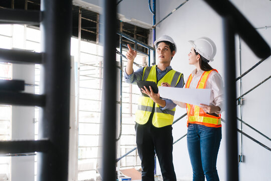 Asian male and female engineers wearing safety vests and helmets inspect structural elements, review blueprints, plan material choices bricks, steel, concrete during home renovation project on-site.
