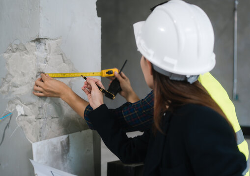Asian male and female engineers wearing safety vests and helmets inspect structural elements, review blueprints, plan material choices bricks, steel, concrete during home renovation project on-site.