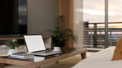 Blank screen laptop on glass top wooden table in a room with city view terrace behind the glass door