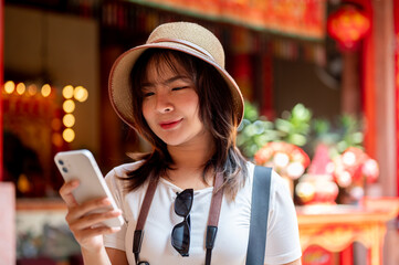 Asian woman wearing hat looking at her smartphone after taking photos in the Chinese shrine temple.