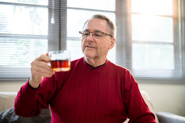 Old white caucasian man wearing glasses looking pleased with his coffee sitting on living room sofa.