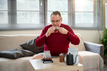Glasses old white caucasian man smelling coffee aroma from his cup sitting on sofa in a living room.