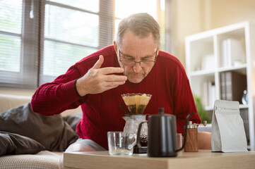 Old white caucasian man wearing glasses smelling coffee aroma from filter paper on dripper at table.