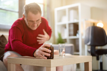 Old white caucasian man is getting a heart attack or cardiac arrest sitting on sofa in a living room
