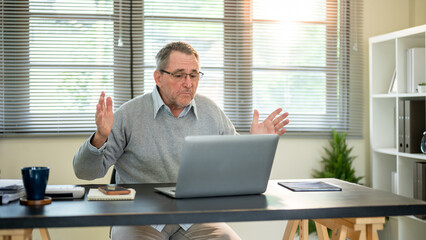 Old white caucasian man wearing glasses looking worried at laptop with arms raising at working table