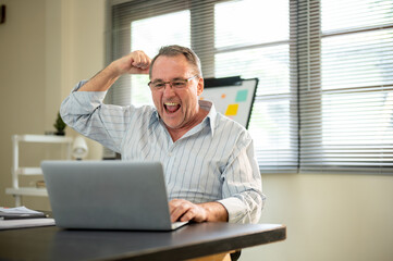 Old white caucasian man with glasses raising a fist while looking at laptop on office working table.