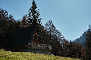 A modern wooden A-frame cabin with a stone foundation stands under a clear blue sky, surrounded by autumn forest in the Montenegro mountains, offering a cozy retreat in nature.