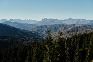 A scenic autumn landscape in the Komovi mountains of Montenegro, featuring layers of forested hills and distant rocky peaks under a clear blue sky.