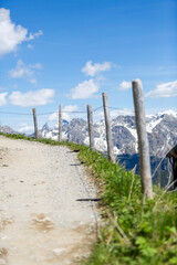 Schmaler Weg mit Holzpfosten am Rand, führt durch eine alpine Landschaft mit schneebedeckten Bergen