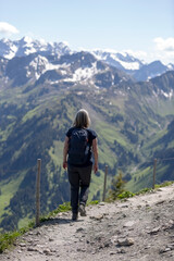 Eine Wanderin auf einem Bergpfad &uuml;ber gr&uuml;ne H&auml;nge mit Blick in die weite, schneebedeckte Berglandschaft der Alpen