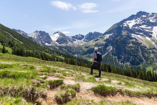 Eine Wanderin auf einem Bergpfad über grüne Hänge mit Blick in die weite, schneebedeckte Berglandschaft der Alpen