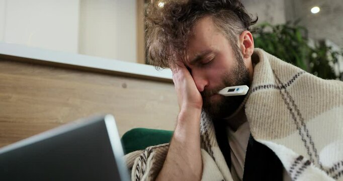 Close up ill male sitting on sofa at home office under plaid, working remotely, using paper tissue with termometr in mouth. Man feeling unwell, sick with running nose, need a rest.