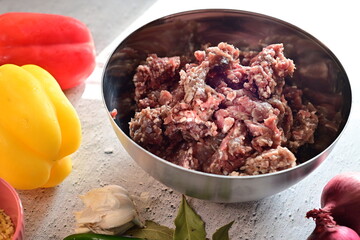 Raw ground beef in a silver bowl surrounded by fresh vegetables on a light background. A clean, bright setup representing home cooking, meal preparation, and fresh ingredients